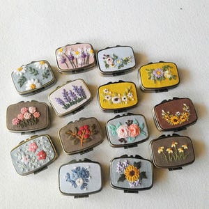Pode incluir: Uma coleção de pequenas caixas retangulares bordadas com desenhos florais. As caixas apresentam várias cores e tipos de flores, incluindo margaridas, lavanda e girassóis. Cada caixa tem uma moldura de metal e uma tampa articulada.