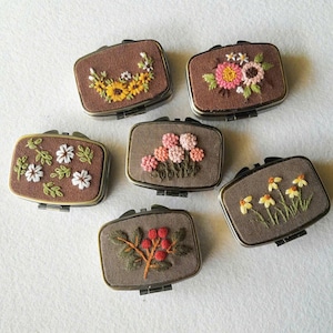 Puede incluir: Una colección de seis pequeñas cajas rectangulares marrones con tapas con bisagras. Cada caja presenta intrincados bordados florales en varios colores, incluyendo girasoles amarillos, flores rosas y blancas, y hojas verdes. Las cajas tienen un borde metálico.