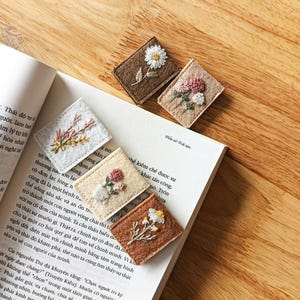 Può includere: Una collezione di segnalibri in feltro ricamati con disegni floreali. I segnalibri rettangolari sono disponibili in vari colori come marrone, beige e bianco. Ogni segnalibro mostra un fiore diverso, tra cui margherite, con cuciture dettagliate.