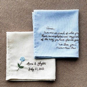 Peut inclure: Deux mouchoirs brodés. L'un est bleu clair avec un message manuscrit en fil noir. L'autre est blanc cassé avec une fleur bleue et les noms "Anna & Skylar" et la date "July 27, 2025" brodés en fil noir.