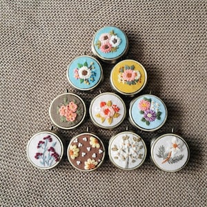 Peut inclure: Dix petits boîtiers à bijoux ronds brodés, chacun avec un motif floral différent. Les boîtes présentent une variété de couleurs, dont le rose, le jaune, le bleu et le blanc. Elles ont un bord doré et sont disposées sur une surface texturée de ton neutre.