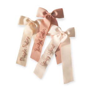 K&ouml;nnte beinhalten: Drei rosa und cremefarbene Satinb&auml;nder mit personalisierten Namen, die in goldfarbener Stickerei ausgef&uuml;hrt sind. Die Namen sind "Maple Faline", "Rachel Cutler" und "Daisy Ward".