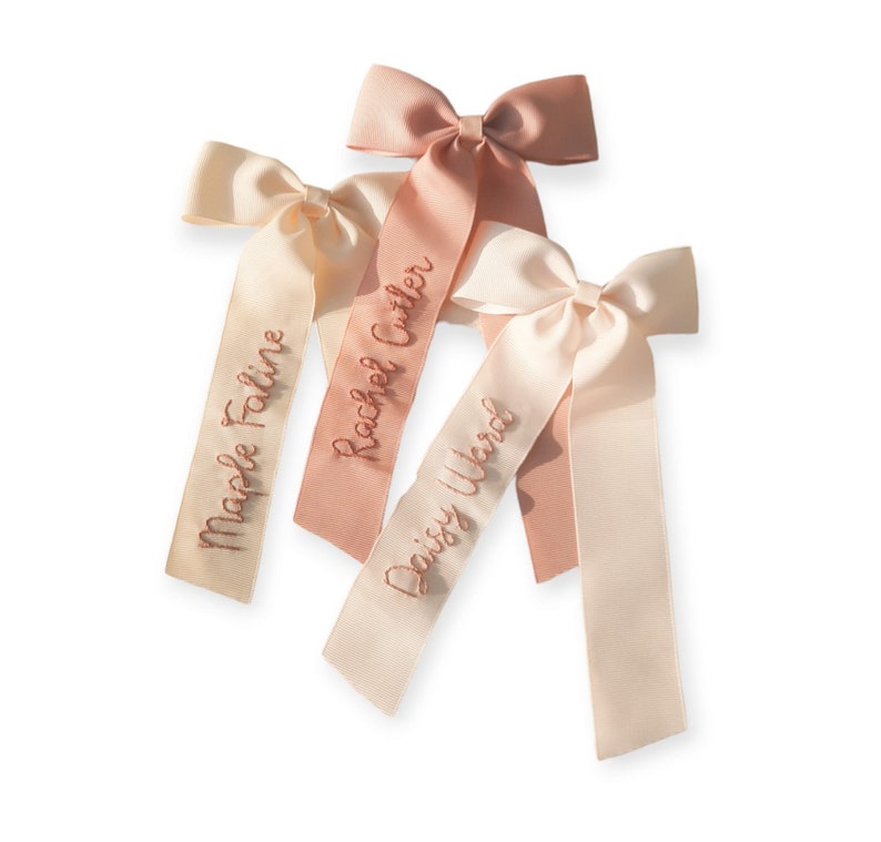 K&ouml;nnte beinhalten: Drei rosa und cremefarbene Satinb&auml;nder mit personalisierten Namen, die in goldfarbener Stickerei ausgef&uuml;hrt sind. Die Namen sind "Maple Faline", "Rachel Cutler" und "Daisy Ward".