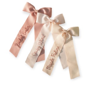 K&ouml;nnte beinhalten: Drei rosa und cremefarbene Satin-Haarspangen mit personalisierten Namen, die in rosa gestickt sind: Rachel Cutler, Daisy Ward und Maple Faline.
