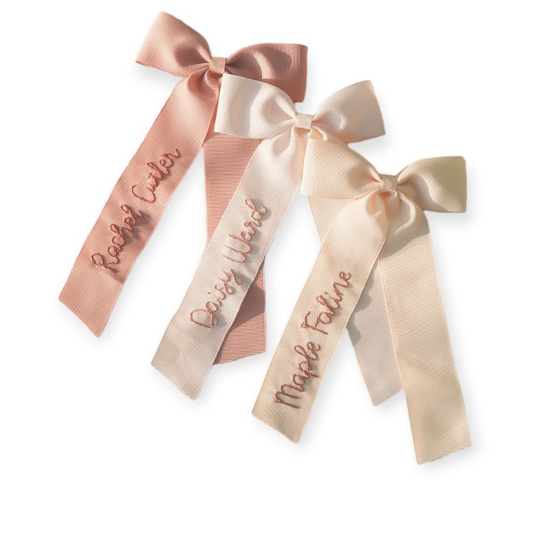 K&ouml;nnte beinhalten: Drei rosa und cremefarbene Satin-Haarspangen mit personalisierten Namen, die in rosa gestickt sind: Rachel Cutler, Daisy Ward und Maple Faline.