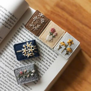 Könnte beinhalten: Eine Sammlung handgefertigter, bestickter Filz-Lesezeichen. Jedes Lesezeichen zeigt ein anderes Blumendesign in verschiedenen Farben, darunter Weiß, Rosa und Gelb, auf einem rechteckigen Hintergrund. Die Lesezeichen liegen auf einem offenen Buch.