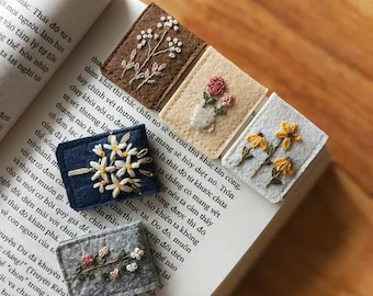 May include: Four rectangular felt bookmarks featuring hand-embroidered floral designs. The bookmarks are brown, cream, navy blue, and gray, each displaying unique botanical patterns including white flowers, pink blossoms, and yellow wildflowers.