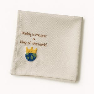 May include: A beige cloth with embroidered text that reads "Daddy is master & King of the world." A small, colorful embroidered image of a crown atop a globe is below the text. The cloth is folded.