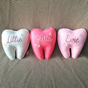 May include: Three heart-shaped tooth pillows in white, pink, and light pink. Each pillow has a name embroidered on it: "Lillie", "Stella", and "Evie". Small flower details are also embroidered on each pillow.