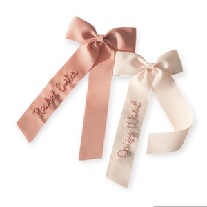 K&ouml;nnte beinhalten: Zwei personalisierte Haarb&auml;nder mit langen Schleifen. Das Band links ist rosa mit dem Namen "Rachel Cutler" bestickt. Das Band rechts ist wei&szlig; mit dem Namen "Daisy Ward" bestickt.