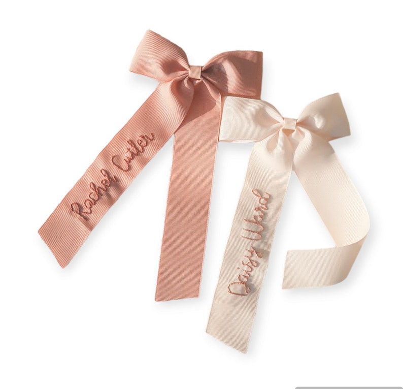 K&ouml;nnte beinhalten: Zwei personalisierte Haarb&auml;nder mit langen Schleifen. Das Band links ist rosa mit dem Namen "Rachel Cutler" bestickt. Das Band rechts ist wei&szlig; mit dem Namen "Daisy Ward" bestickt.