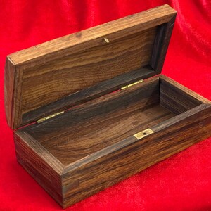 May include: An open, rectangular wooden box with a hinged lid. The box is made of dark brown wood with visible grain patterns. The interior is lined with a darker wood. Brass-colored hinges and a clasp secure the lid. The box sits on a red surface.