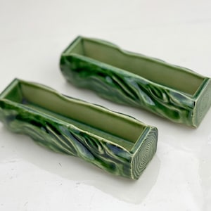 May include: Two green ceramic planters, log-shaped with carved bark and tree ring details. The rectangular openings are visible. The planters are a muted green colour, suitable for small plants or decorative items.