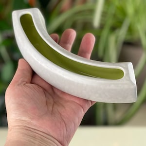 May include: A curved, white ceramic vase with a light green interior. The vase has a rectangular shape with rounded edges and a long, open trough. The vase is held in a hand.