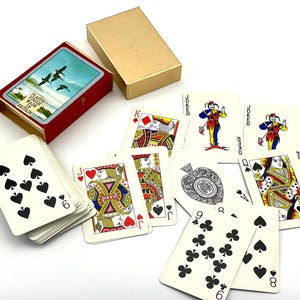 Könnte beinhalten: Ein Kartenspiel in einer roten und goldenen Schachtel. Die Schachtel zeigt ein Bild von fliegenden Vögeln. Mehrere Karten sind sichtbar, darunter ein König, eine Königin, ein Bube und Joker. Die Karten haben einen weißen Hintergrund mit schwarzen und roten Farben.