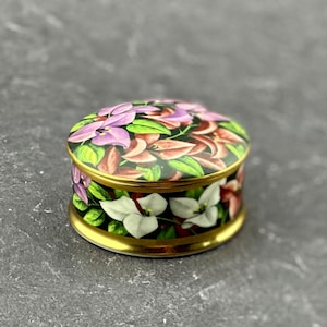 May include: A small, round trinket box with a floral design. The box is white with a gold trim and features pink, purple, and white flowers on a black background.