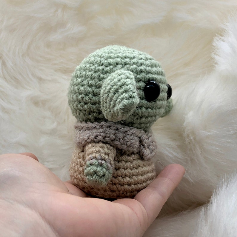 Puede incluir: Mu&ntilde;eco de ganchillo que se asemeja a un personaje con grandes ojos negros y orejas grandes. El mu&ntilde;eco es principalmente verde claro con un atuendo beige y gris. El mu&ntilde;eco se sostiene en una mano sobre un fondo blanco y esponjoso.
