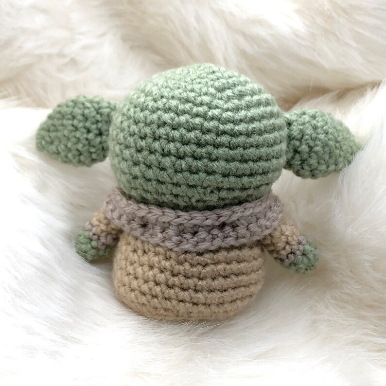 Puede incluir: Una figura de ganchillo de un personaje con orejas grandes, en tonos verdes, beige y grises. La mu&ntilde;eca hecha a mano tiene una cabeza redonda, orejas grandes y un cuerpo peque&ntilde;o. La figura est&aacute; sobre una superficie blanca y esponjosa.