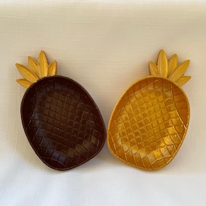 May include: Two pineapple-shaped decorative trays. One is dark brown, the other is gold. Both have a textured surface and a gold-coloured crown. These trays are ideal for holding small items.