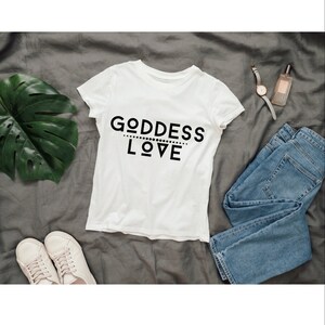 May include: White t-shirt with black text that reads "GODDESS LOVE" on a gray background. The shirt is laid flat with a pair of white sneakers, blue jeans, a watch, and a bottle of perfume.