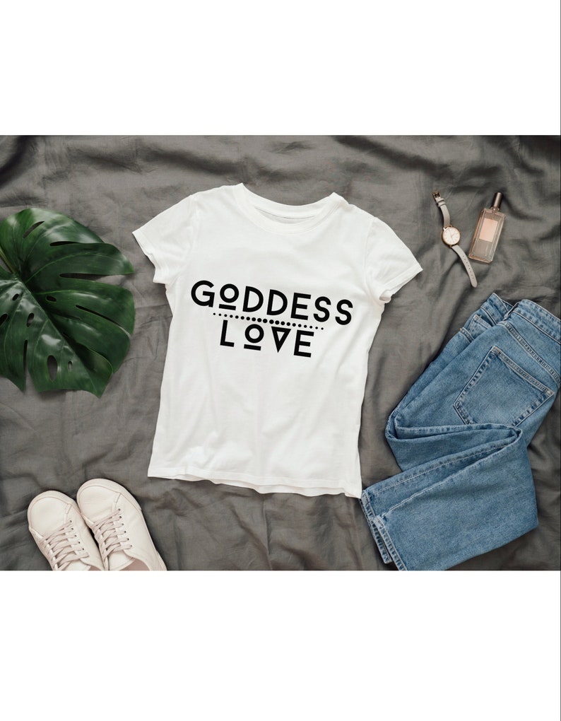 May include: White t-shirt with black text that reads "GODDESS LOVE" on a gray background. The shirt is laid flat with a pair of white sneakers, blue jeans, a watch, and a bottle of perfume.