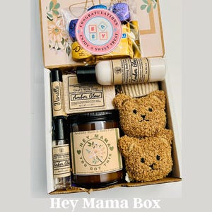 May include: A gift box filled with self-care items. Includes a candle, soap, body lotion, lip balm, and a rollerball perfume. The box also contains two teddy bear-shaped washcloths and a package of candies. The box is labeled "Hey Mama Box".