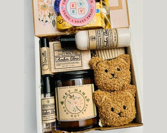 May include: A gift box filled with self-care items. Includes a candle, soap, body lotion, lip balm, and a rollerball perfume. The box also contains two teddy bear-shaped washcloths and a package of candies. The box is labeled "Hey Mama Box".
