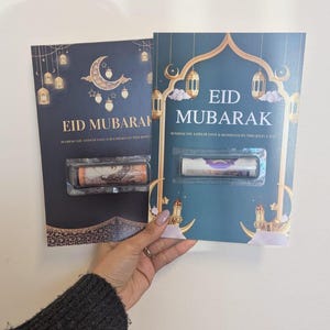 Puede incluir: Dos tarjetas de felicitaci&oacute;n color teal con detalles dorados, con el texto "EID MUBARAK". Las tarjetas est&aacute;n decoradas con faroles y lunas crecientes. Cada tarjeta contiene un art&iacute;culo enrollado en una funda de pl&aacute;stico transparente.