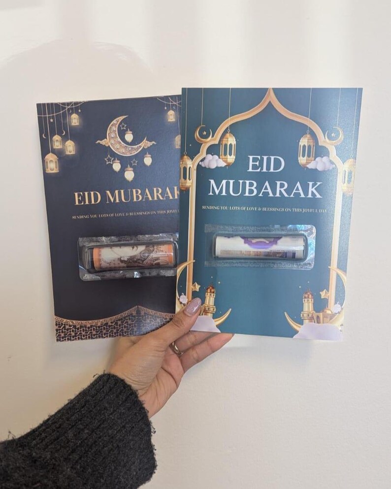 Puede incluir: Dos tarjetas de felicitaci&oacute;n color teal con detalles dorados, con el texto "EID MUBARAK". Las tarjetas est&aacute;n decoradas con faroles y lunas crecientes. Cada tarjeta contiene un art&iacute;culo enrollado en una funda de pl&aacute;stico transparente.