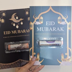 Puede incluir: Dos tarjetas de felicitaci&oacute;n de Eid Mubarak. Una tarjeta es negra con detalles dorados, una luna creciente y faroles. La otra es verde azulado con detalles dorados, faroles y un dise&ntilde;o de mezquita. Cada tarjeta contiene un art&iacute;culo enrollado en una funda de pl&aacute;stico transparente.