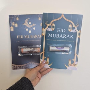 Puede incluir: Dos tarjetas de felicitaci&oacute;n Eid Mubarak azul marino con detalles dorados, una luna creciente y faroles. Cada tarjeta tiene una funda de pl&aacute;stico transparente que contiene billetes enrollados. El texto "EID MUBARAK" se muestra.
