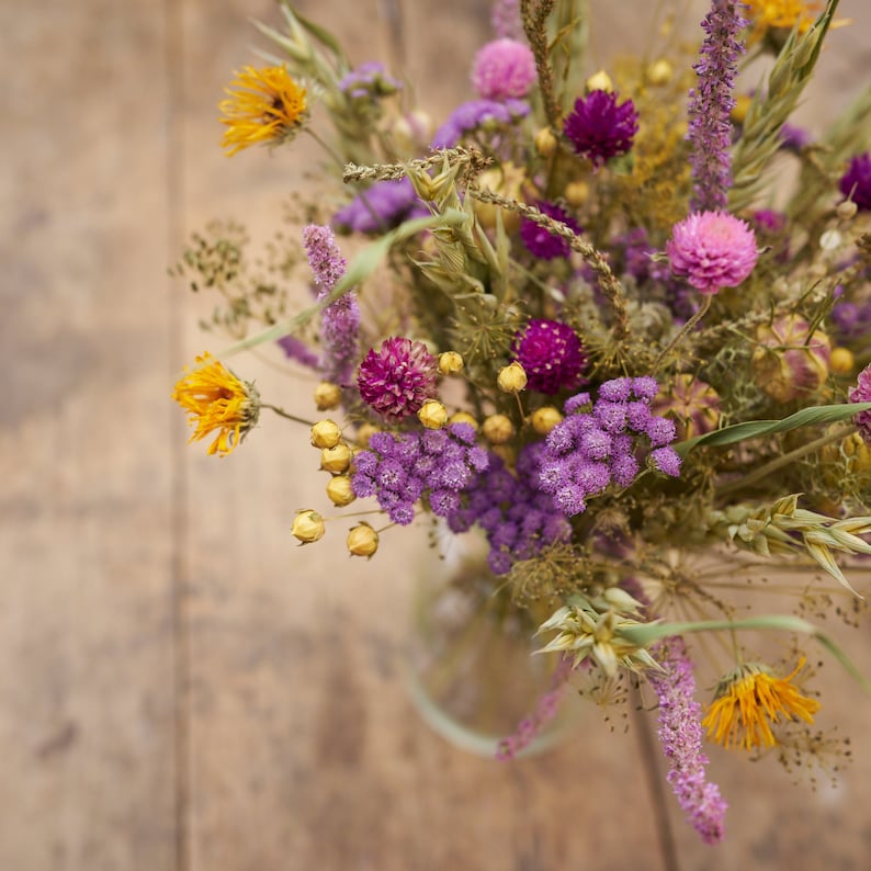 May include: A bouquet of dried flowers in a glass vase. The flowers are a mix of purple, yellow, and white. The bouquet is sitting on a wooden table.