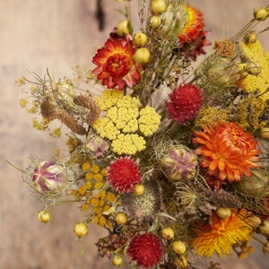 May include: A dried flower arrangement with a variety of flowers in shades of yellow, orange, and red. The flowers are arranged in a vase and are surrounded by dried grasses and leaves.
