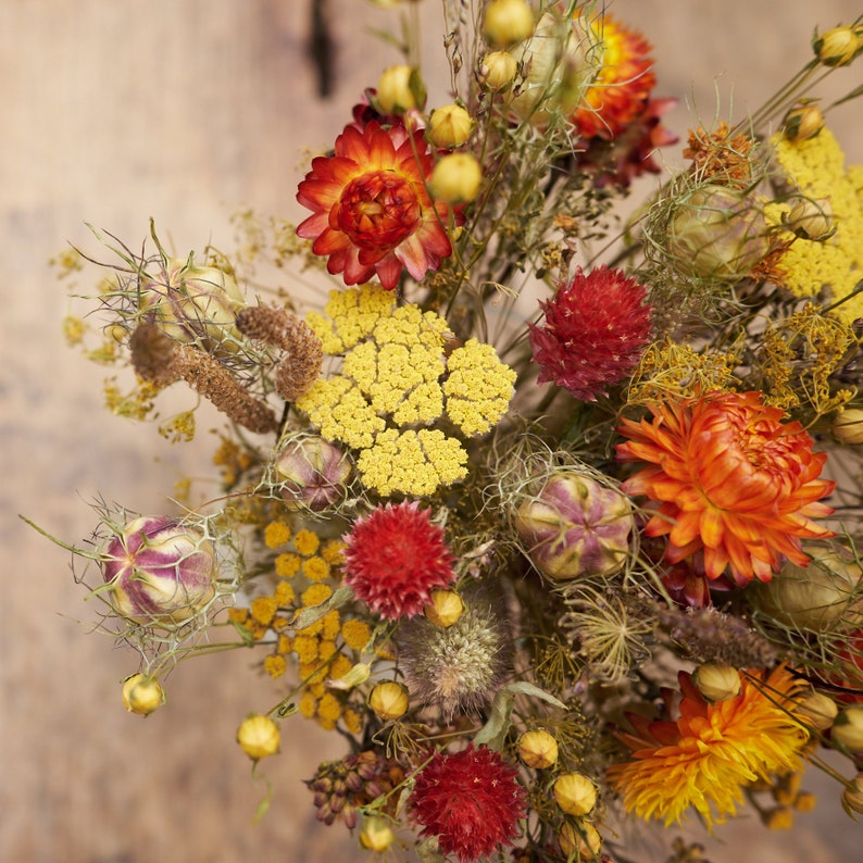 May include: A dried flower arrangement with a variety of flowers in shades of yellow, orange, and red. The flowers are arranged in a vase and are surrounded by dried grasses and leaves.