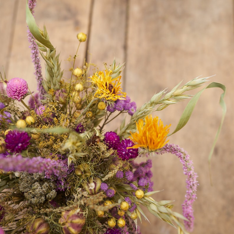 May include: A dried flower bouquet with purple, yellow, and orange flowers. The bouquet is arranged in a vase and is sitting on a wooden surface.