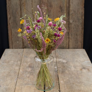 May include: A bouquet of dried flowers in a clear glass vase. The flowers are a mix of purple, pink, yellow, and green. The vase is on a wooden table.