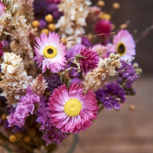May include: A bouquet of dried flowers in shades of pink, purple, and white. The bouquet includes dried daisies, lavender, and other wildflowers.