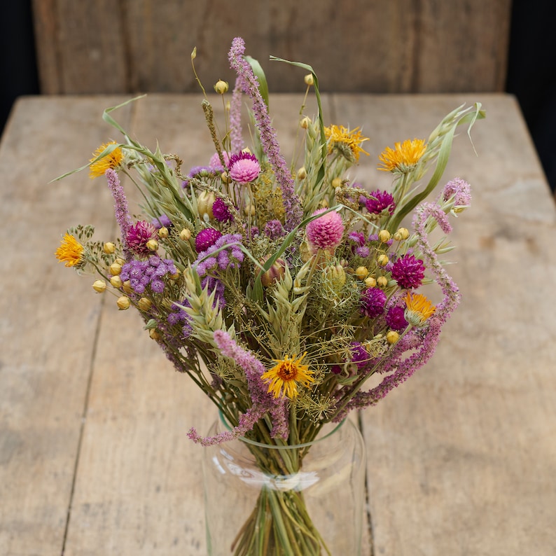 May include: A bouquet of dried flowers in a clear glass vase. The bouquet includes purple, pink, and yellow flowers, as well as dried grasses.