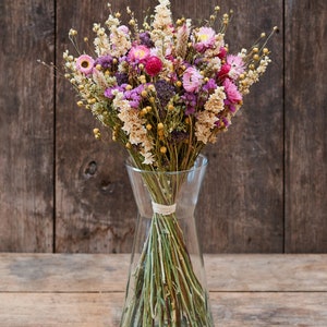 May include: A bouquet of dried flowers in shades of pink, purple, and cream, arranged in a clear glass vase. The flowers are tied together with a natural twine.