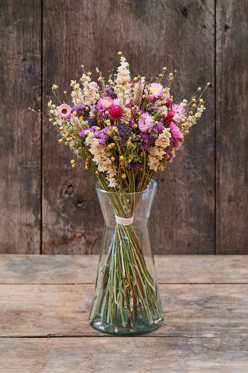 May include: A bouquet of dried flowers in shades of pink, purple, and cream, arranged in a clear glass vase. The flowers are tied together with a natural twine.