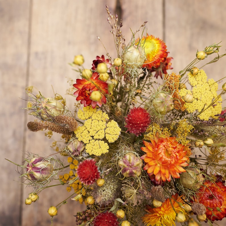 May include: A dried flower arrangement with a variety of colors and textures, including yellow, orange, red, and brown. The flowers are arranged in a vase and are surrounded by greenery.