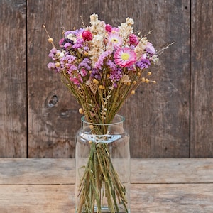 May include: A bouquet of dried flowers in shades of pink, purple, and white, arranged in a clear glass vase. The flowers are set against a rustic wooden background.