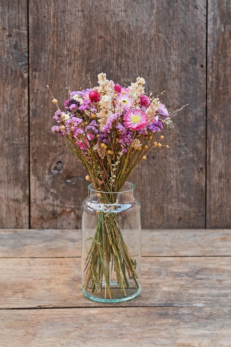May include: A bouquet of dried flowers in shades of pink, purple, and white, arranged in a clear glass vase. The flowers are set against a rustic wooden background.
