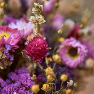 May include: A bouquet of dried flowers in shades of pink, purple, and yellow. The flowers are arranged in a loose, natural style, and the bouquet is tied with a brown ribbon.