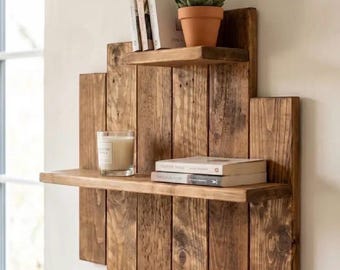 May include: A rustic, three-tiered wooden shelf unit with a natural finish. The shelves hold books, a potted succulent, and a candle in a glass. The unit is mounted on a light-colored wall, creating a warm, inviting display.
