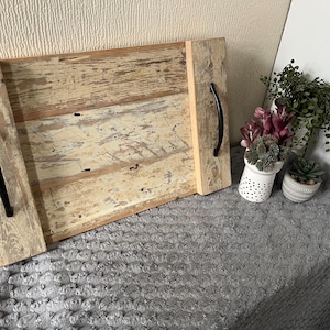 May include: A rectangular wooden serving tray with a distressed finish. The tray has two black handles and is made of light-coloured wood with visible grain and knots. The tray is on a grey surface with decorative plants in the background.