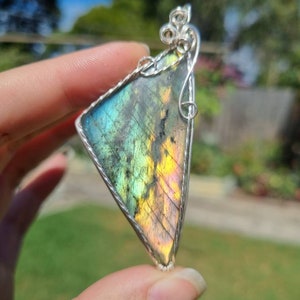 Puede incluir: Un colgante triangular de labradorita con un alambre de plata. La piedra preciosa muestra colores iridiscentes de azul, verde y amarillo. El colgante se sostiene sobre un fondo verde borroso.