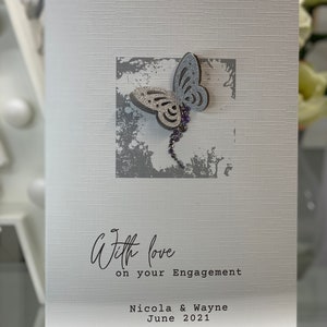 Pu&ograve; includere: Un biglietto bianco con una farfalla argentata glitterata su uno sfondo grigio. Il biglietto dice "With love on your engagement Nicola & Wayne June 2021".