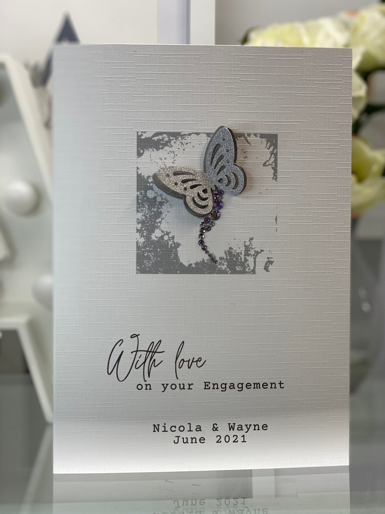 Pu&ograve; includere: Un biglietto bianco con una farfalla argentata glitterata su uno sfondo grigio. Il biglietto dice "With love on your engagement Nicola & Wayne June 2021".