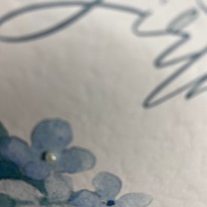 May include: A white background with a blue floral design. The flowers are painted in a watercolor style and have a light blue color. There are small white beads scattered throughout the design.